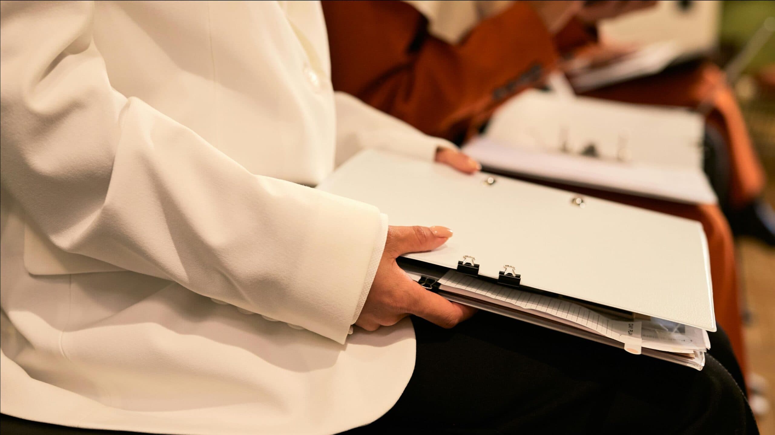 Conference attendee in a white blazer holding a binder of papers on their lap, seated beside other attendees reviewing documents
