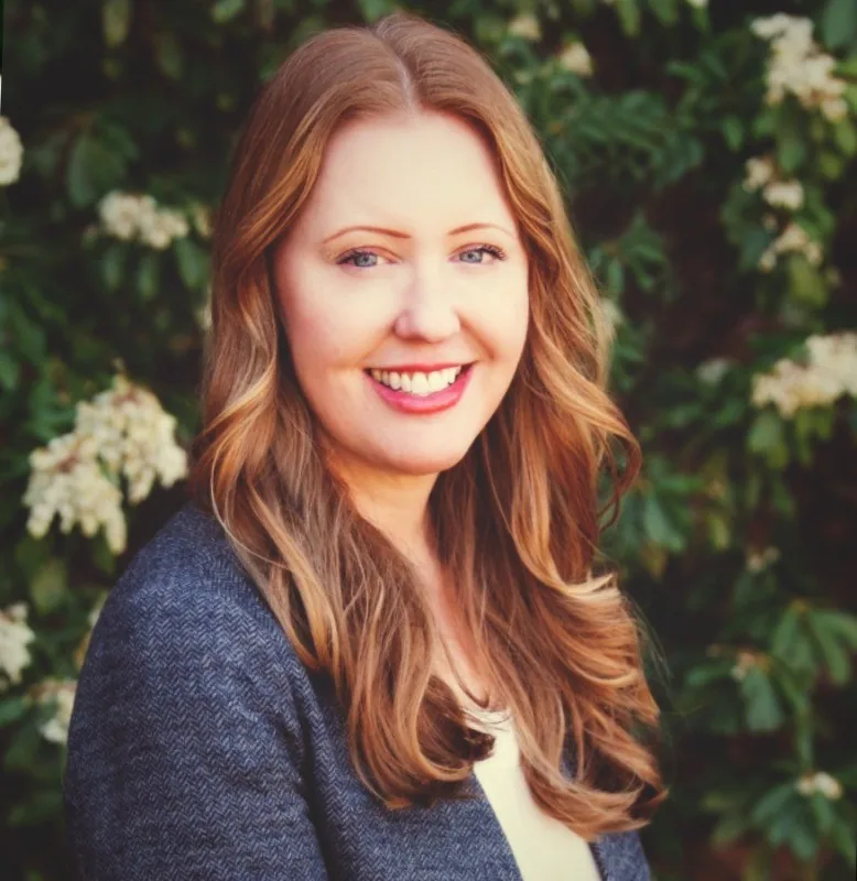 Anna Benyo smiling, with long wavy auburn hair, wearing a grey herringbone blazer over a cream top in front of a flowering green shrub