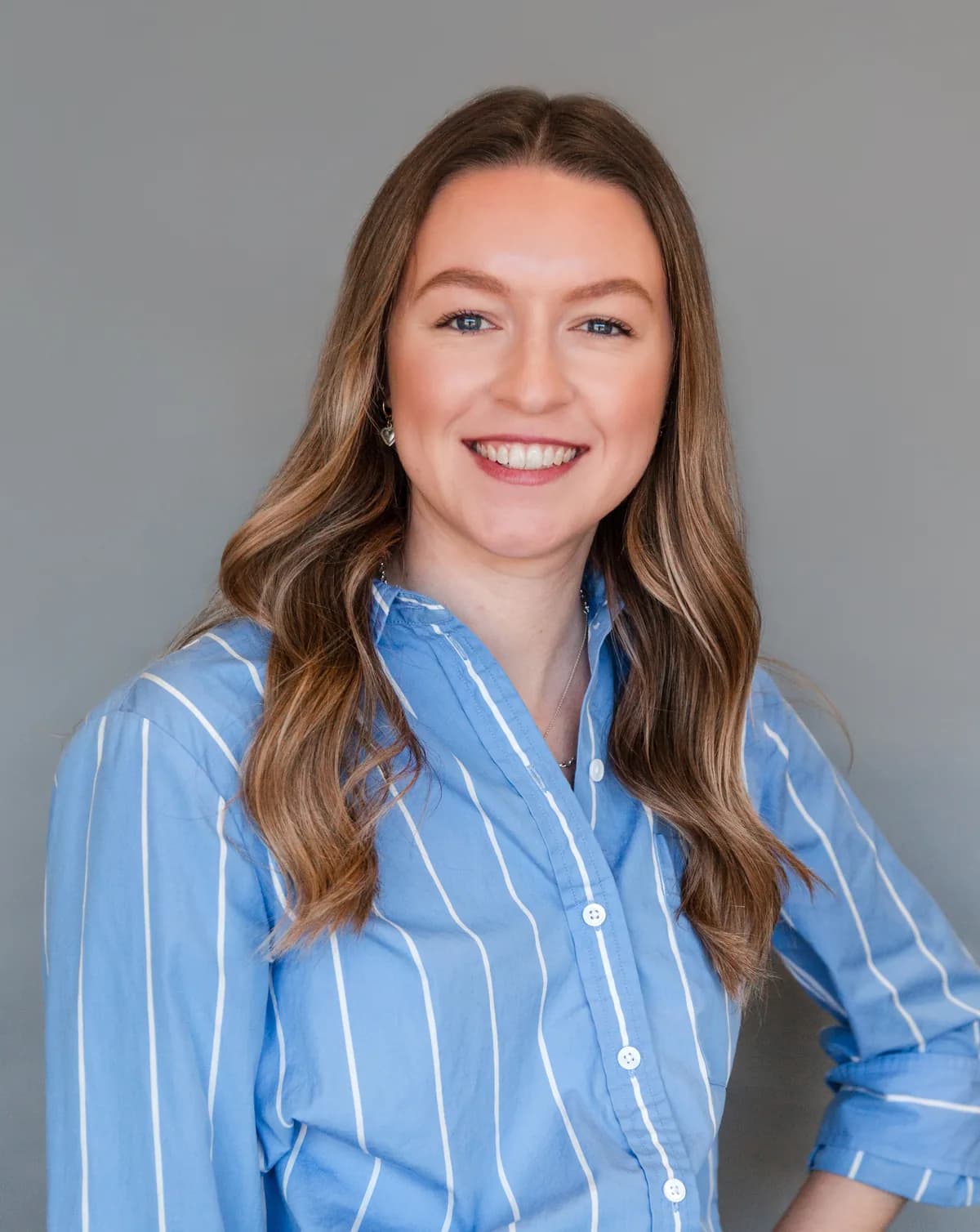 Arden Borders smiling, with long wavy light-brown hair, wearing a blue-and-white striped button-up shirt against a grey backdrop