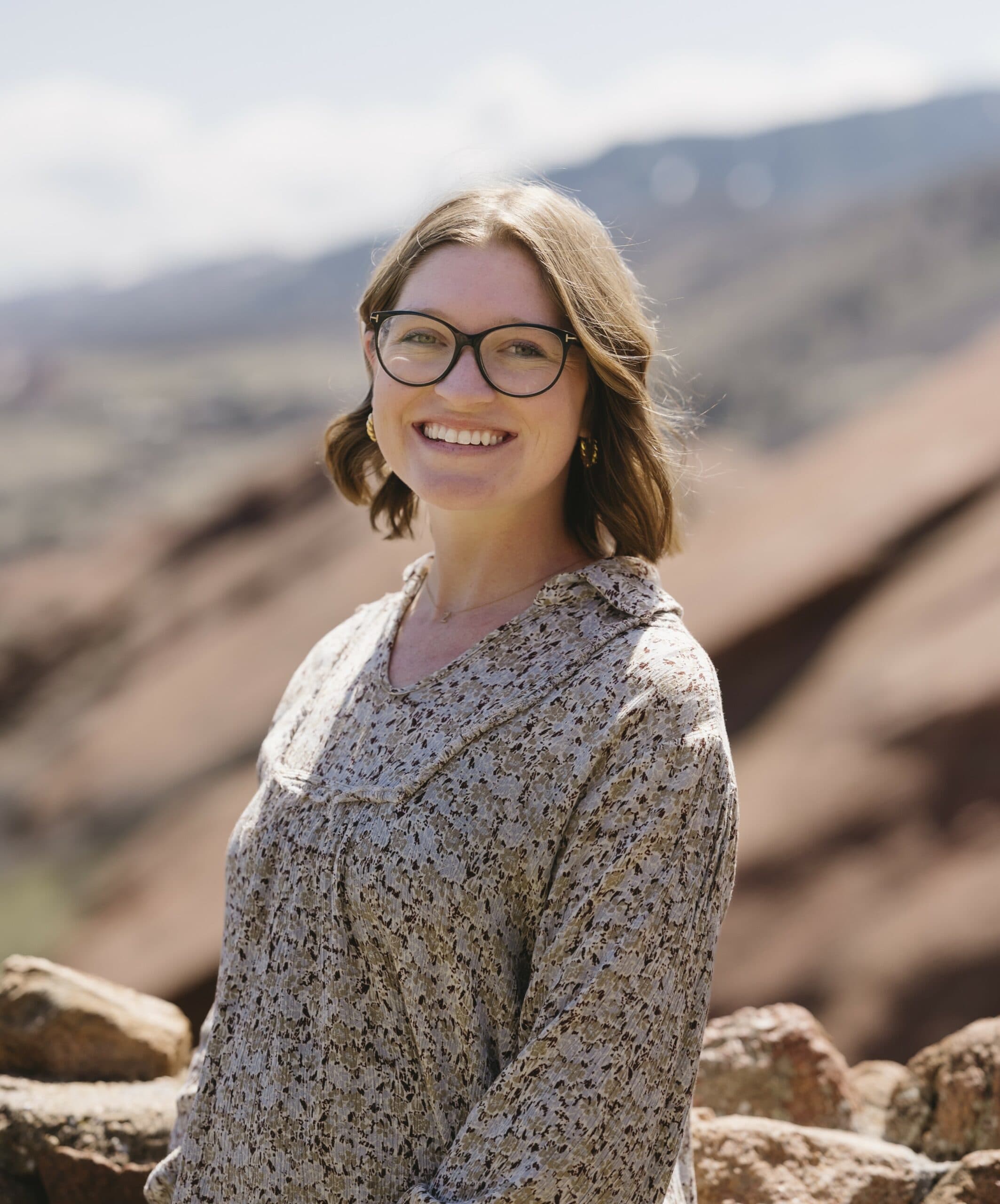 Brianna Robles smiling, with shoulder-length light-brown hair and dark-rimmed glasses, wearing a speckled print blouse in front of blurred red sandstone ridges