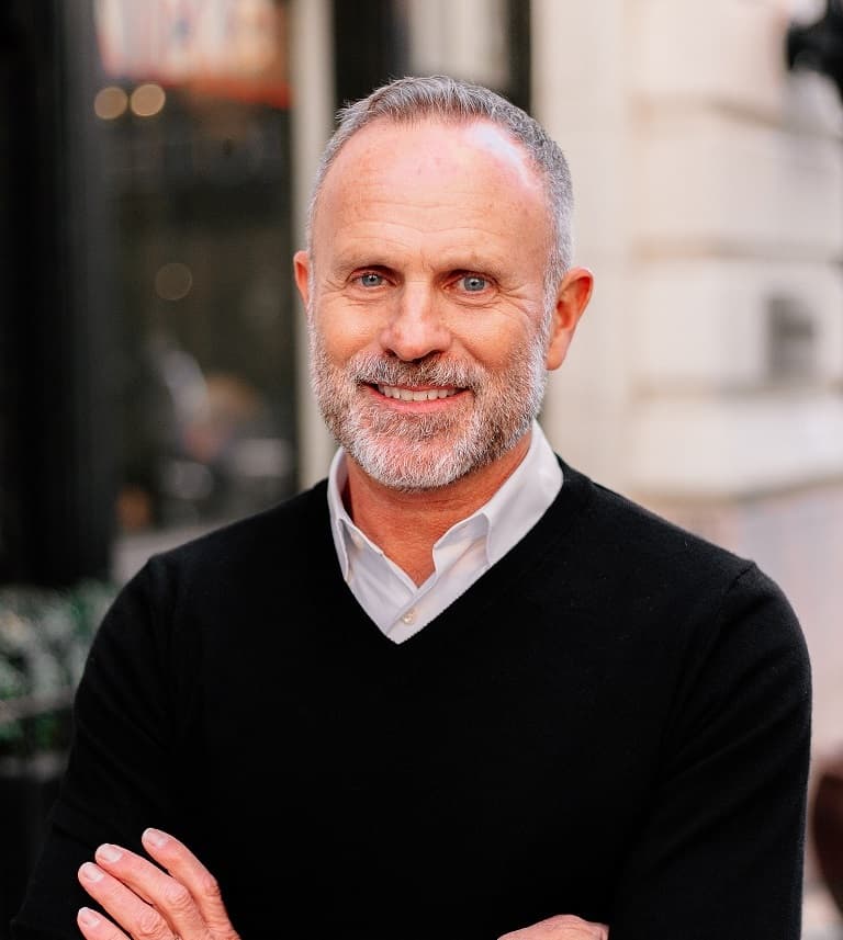 Burke Fishburn smiling, with short grey hair and a grey beard, wearing a black sweater over a collared shirt with arms crossed, on a city street