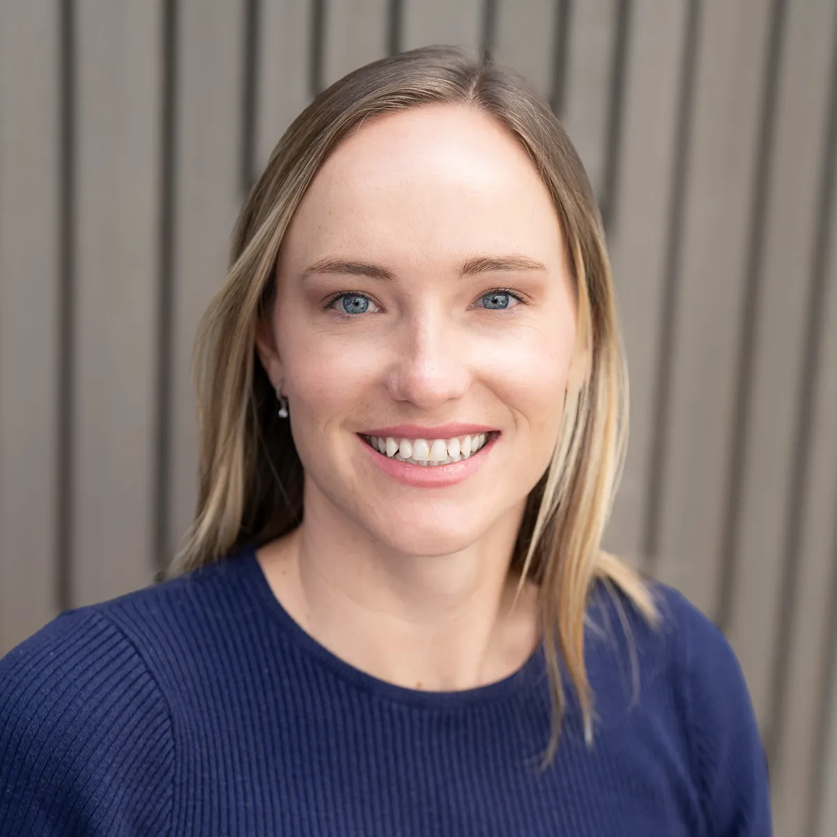 Carina Stavish smiling, with long blonde hair, wearing a navy sweater against a ribbed grey wall