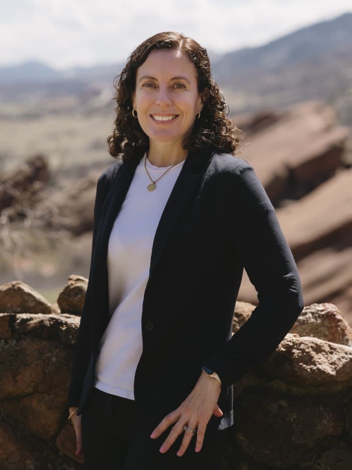 Carla Lewis smiling, with curly dark hair, wearing a black blazer over a white top, leaning on a rock wall with red sandstone ridges behind her