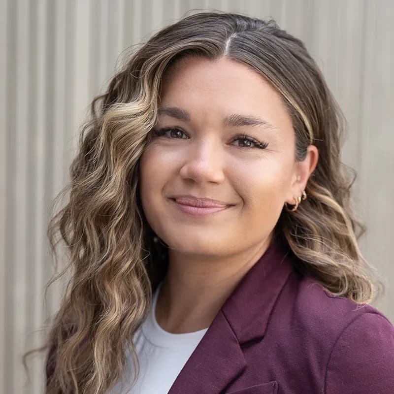 Courtney Lange smiling, with long wavy highlighted hair, wearing a burgundy blazer over a white top against a ribbed grey wall