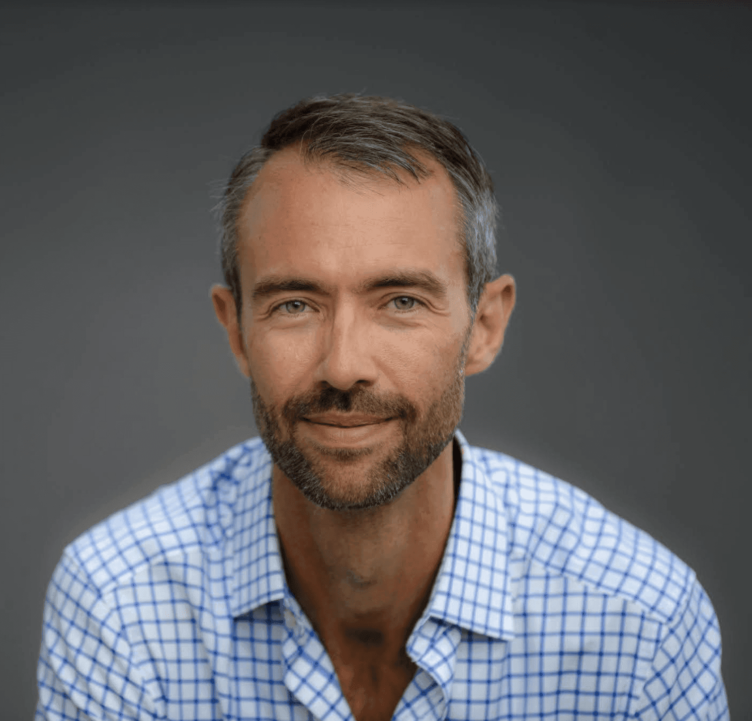 JK Costello smiling, with short dark hair and a beard, wearing a blue checked button-up shirt against a grey backdrop