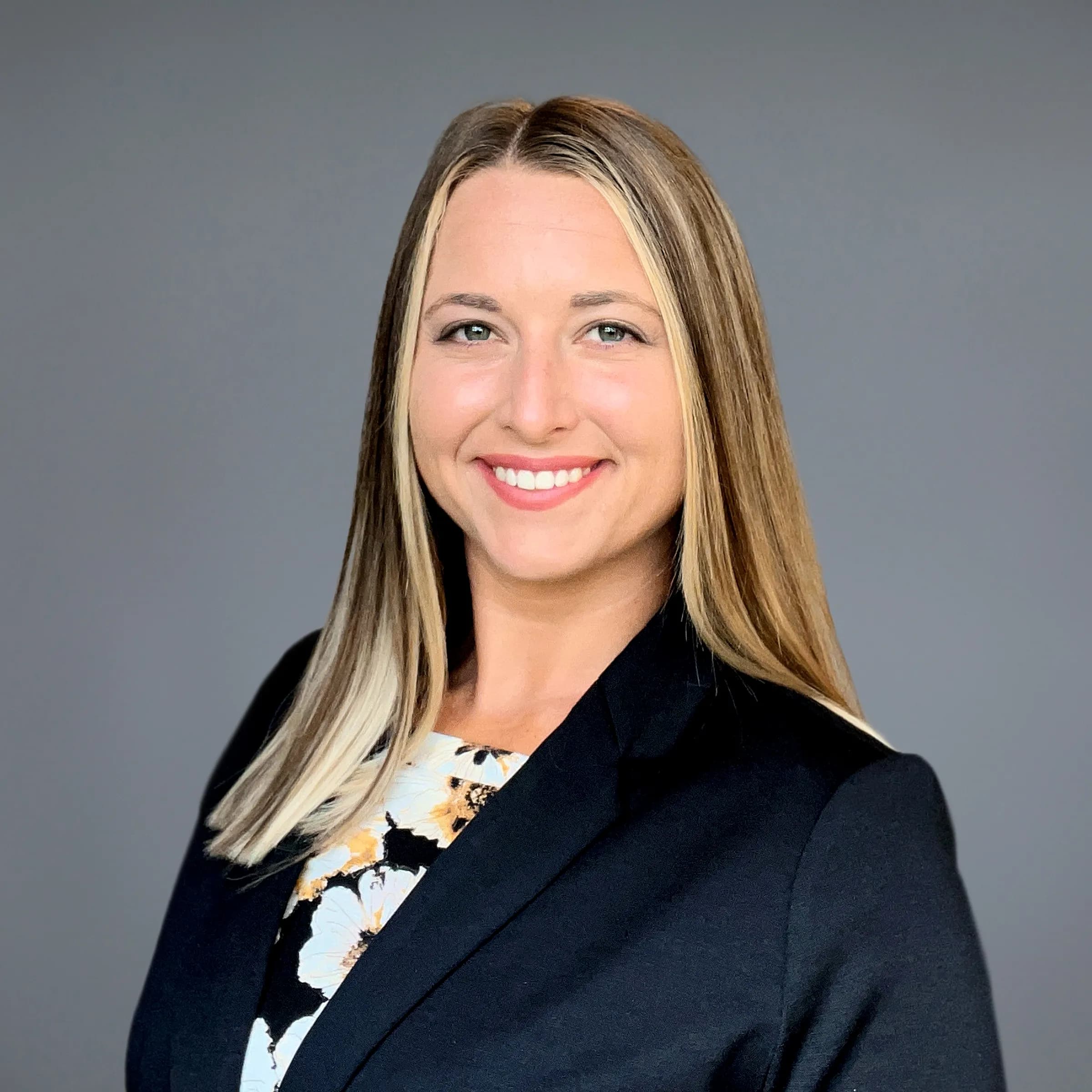Olivia Golan smiling, with long highlighted blonde hair, wearing a black blazer over a floral top against a grey backdrop
