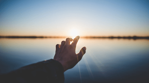 A silhouetted hand reaching toward the setting sun over a calm lake