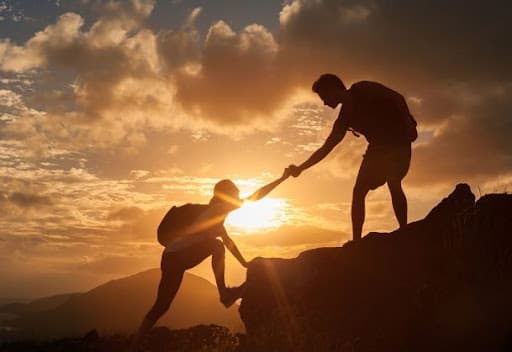Silhouette of one hiker reaching down to help another climb a rocky ridge at sunset