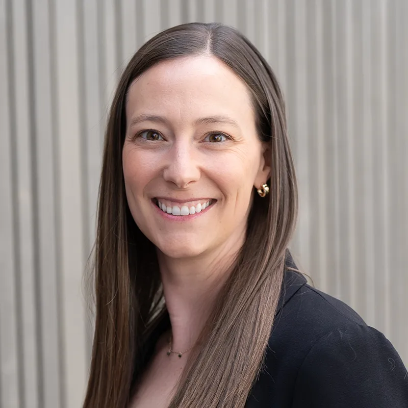 Samantha Strobing smiling, with long straight brown hair, wearing a black top against a ribbed grey wall