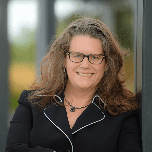 Sharon Steadman smiling, with glasses and long wavy brown hair, wearing a black jacket with white piping, photographed outdoors