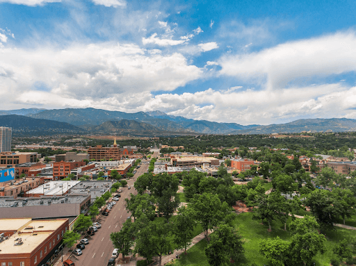 Aerial view of a city's tree-lined main street and a green park, with forested mountains under a partly cloudy blue sky