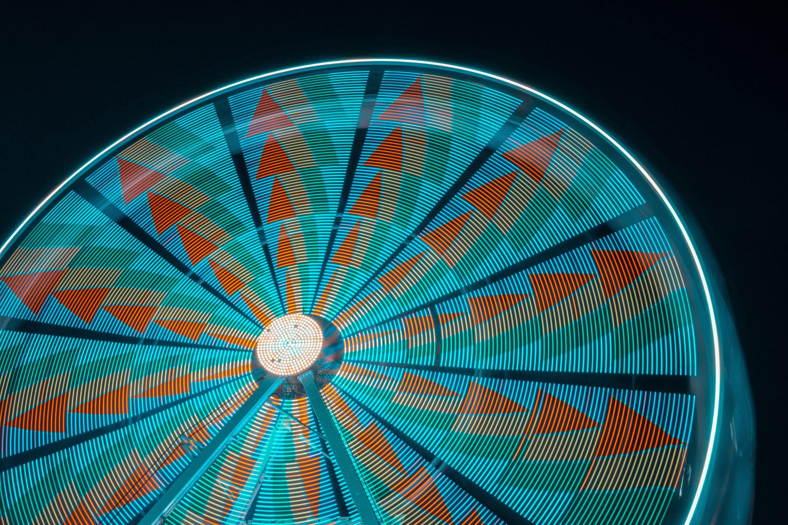 Long-exposure night photo of a spinning Ferris wheel, its spokes blurred into glowing teal rings with orange arrow patterns around a bright hub