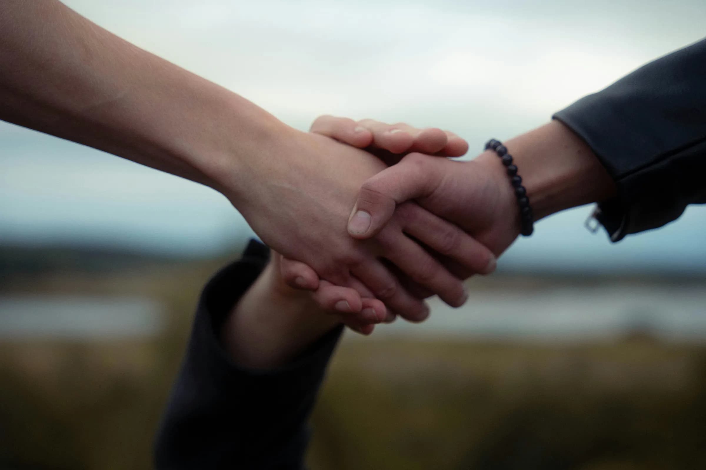 Three people's hands clasped together in a supportive grip, one wrist wearing a dark beaded bracelet, against a blurred outdoor landscape and overcast sky.