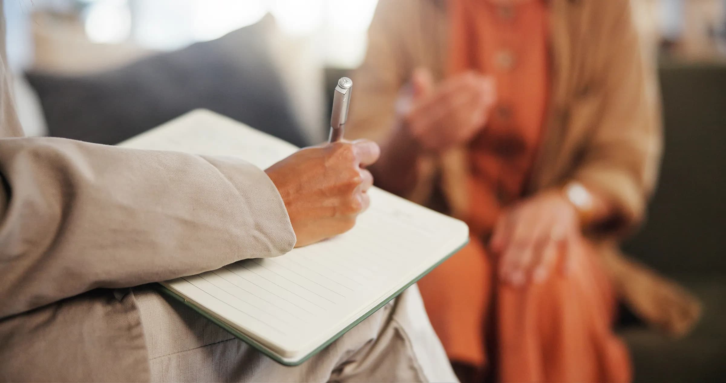 Close-up of a clinician in a beige blazer writing in a notebook during a counseling session, with a client in an orange dress gesturing in the soft-focus background.