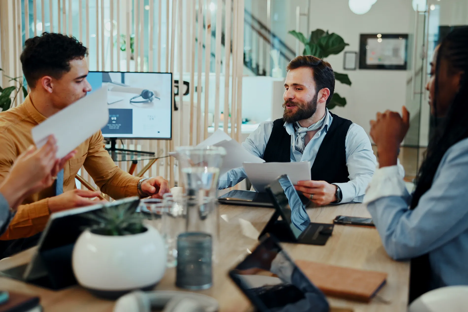 A bearded man in a dark vest and light blue shirt holds papers and talks with colleagues around a conference table with tablets and water glasses in a bright office.