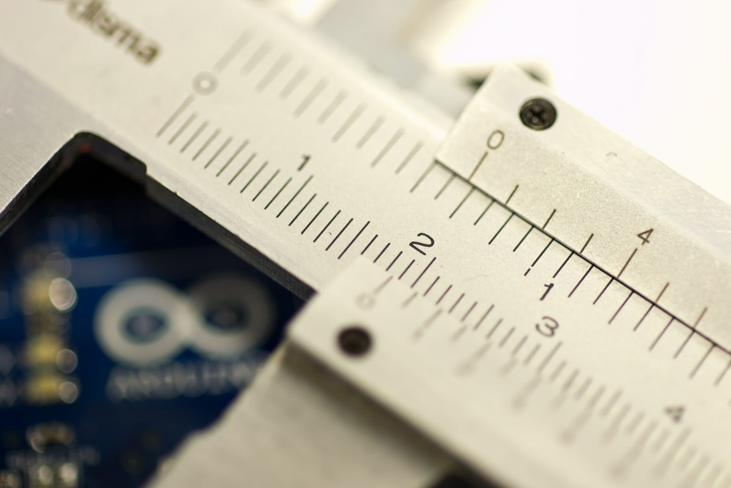 Close-up of the engraved scale on a steel vernier caliper, with a blurred blue circuit board in the background