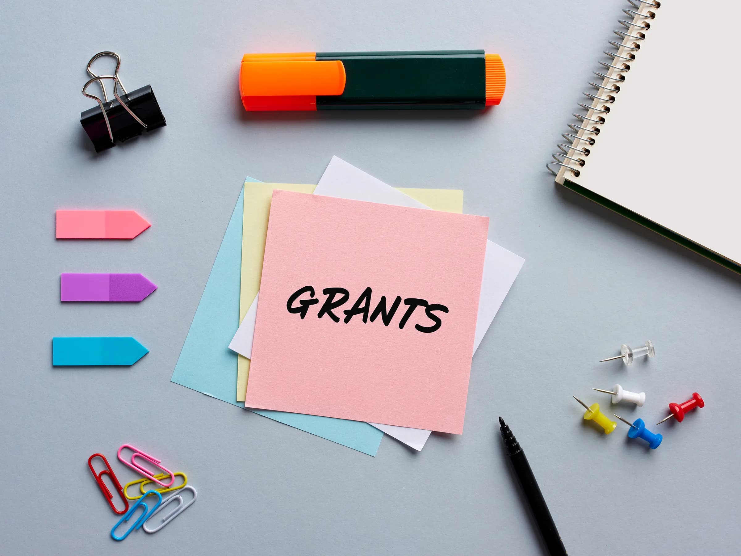 Pink sticky note with the word GRANTS handwritten in black marker, layered over blue, yellow and white notes on a pale blue-grey desk with a highlighter, binder clip, page flags, paper clips, push pins and a spiral notebook