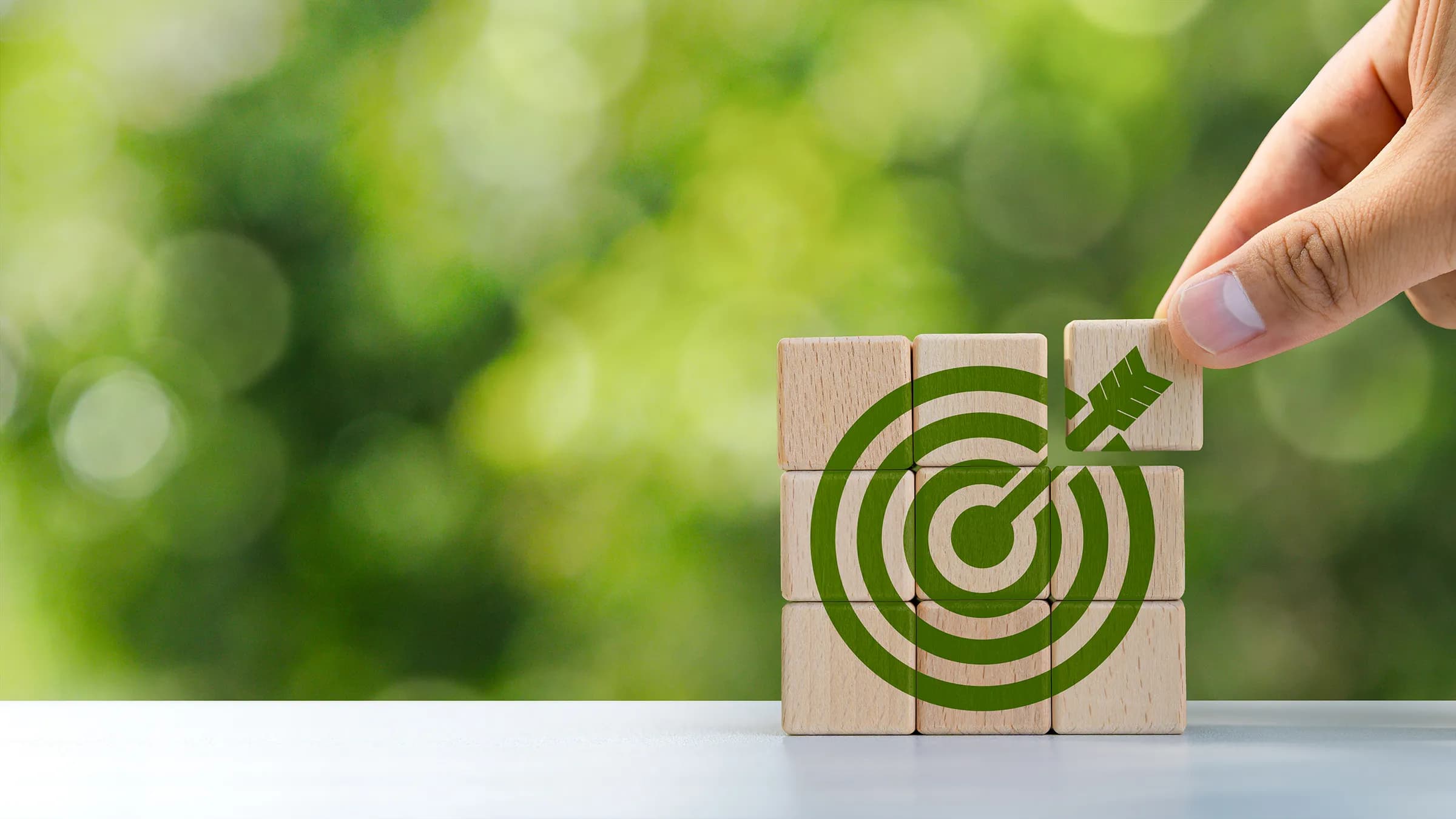 Hand placing the last wooden block, printed with a green arrow, onto a stack of wooden blocks that together form a green bullseye target, against a blurred green background