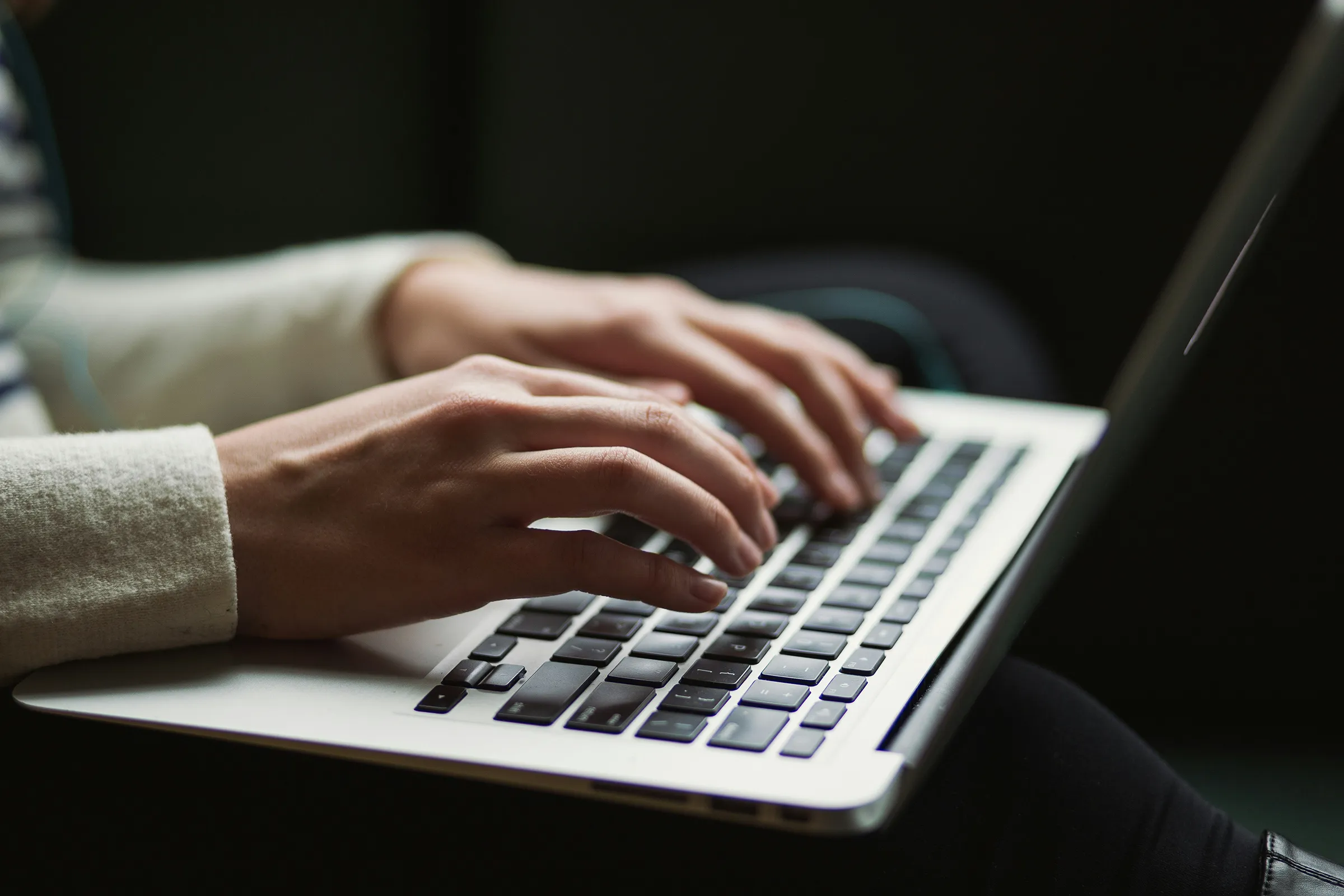 Close-up of hands in a cream knit sweater typing on a silver laptop keyboard against a dark background