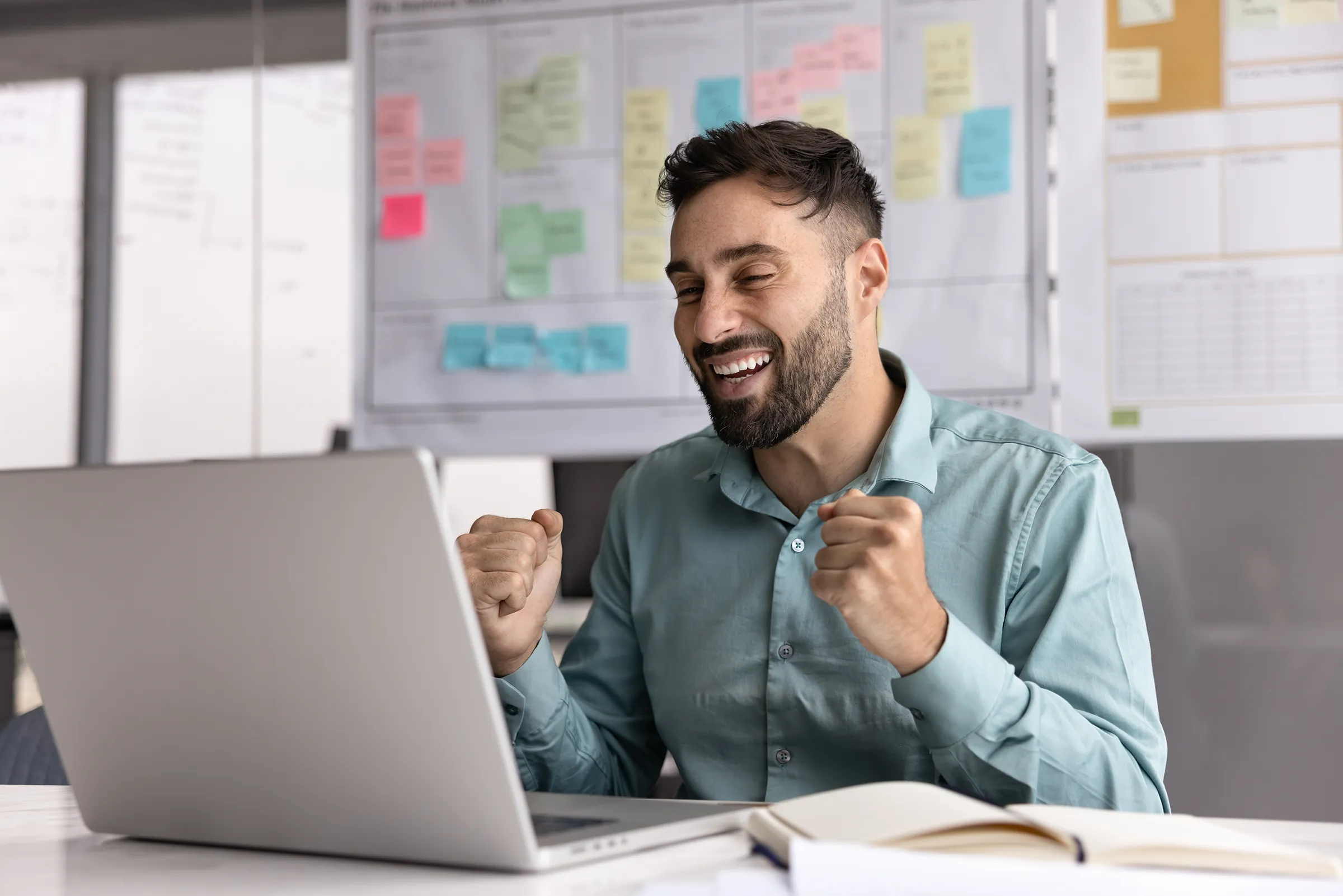 Smiling man in a teal button-down shirt clenching both fists in celebration while looking at a laptop, with a whiteboard of colourful sticky notes behind him