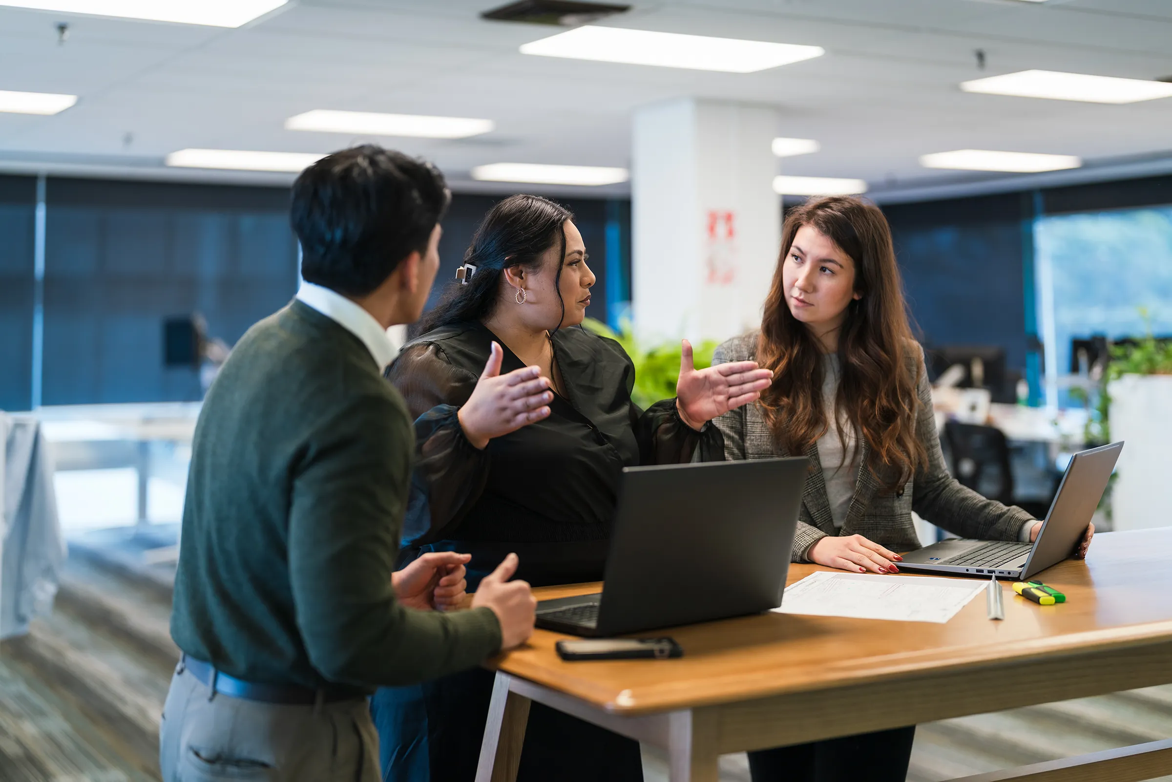 Three colleagues talk around a tall wooden table with open laptops and papers in a bright open-plan office; the woman in the middle gestures with both hands as she explains