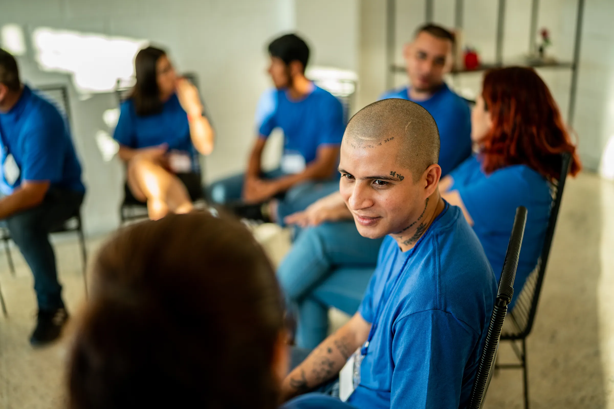 A smiling young man with a shaved head, wearing a blue T-shirt, sits in a circle of chairs during a group session with other participants in matching blue shirts.