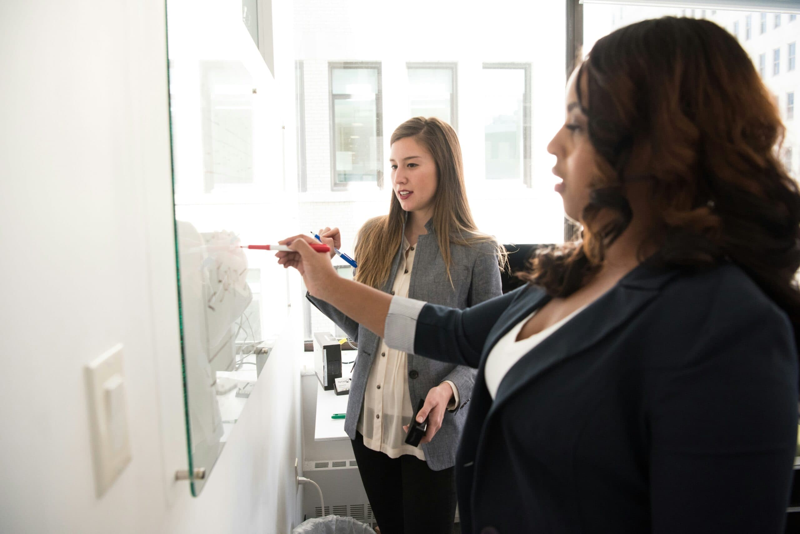 Two women in business attire working at a glass whiteboard in a bright office, one pointing at notes with a red marker