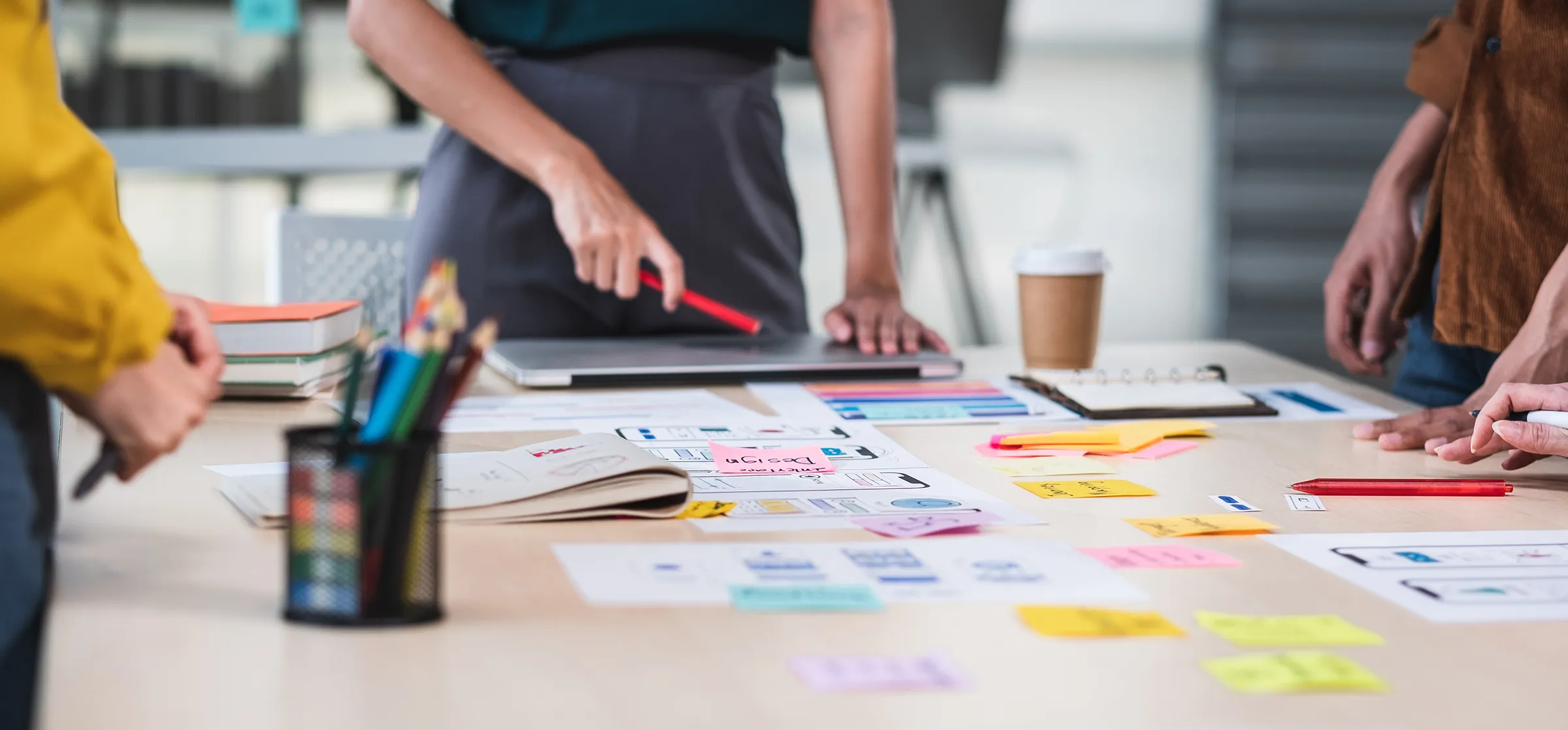 Team members standing around a table covered with wireframe sketches, sticky notes and color charts, one pointing with a red pen