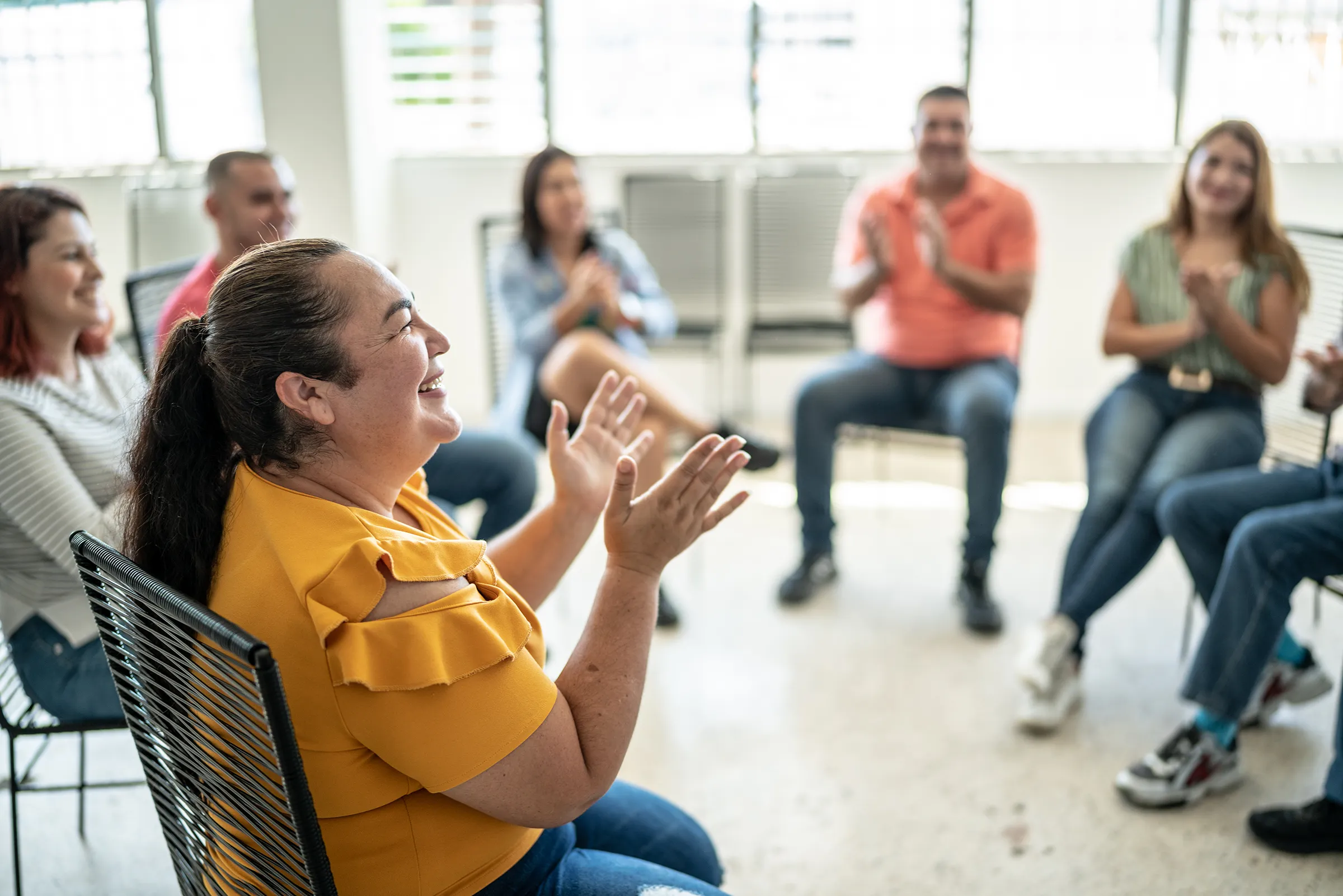 A community support circle in a bright room: a woman in a yellow top laughs and claps in the foreground while others seated in the circle applaud
