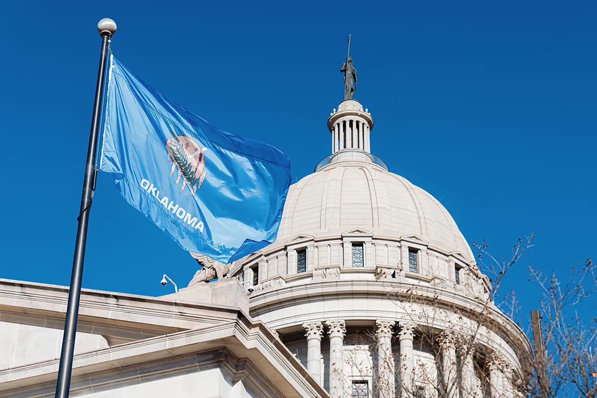 Oklahoma state flag flying beside the domed Oklahoma State Capitol against a clear blue sky