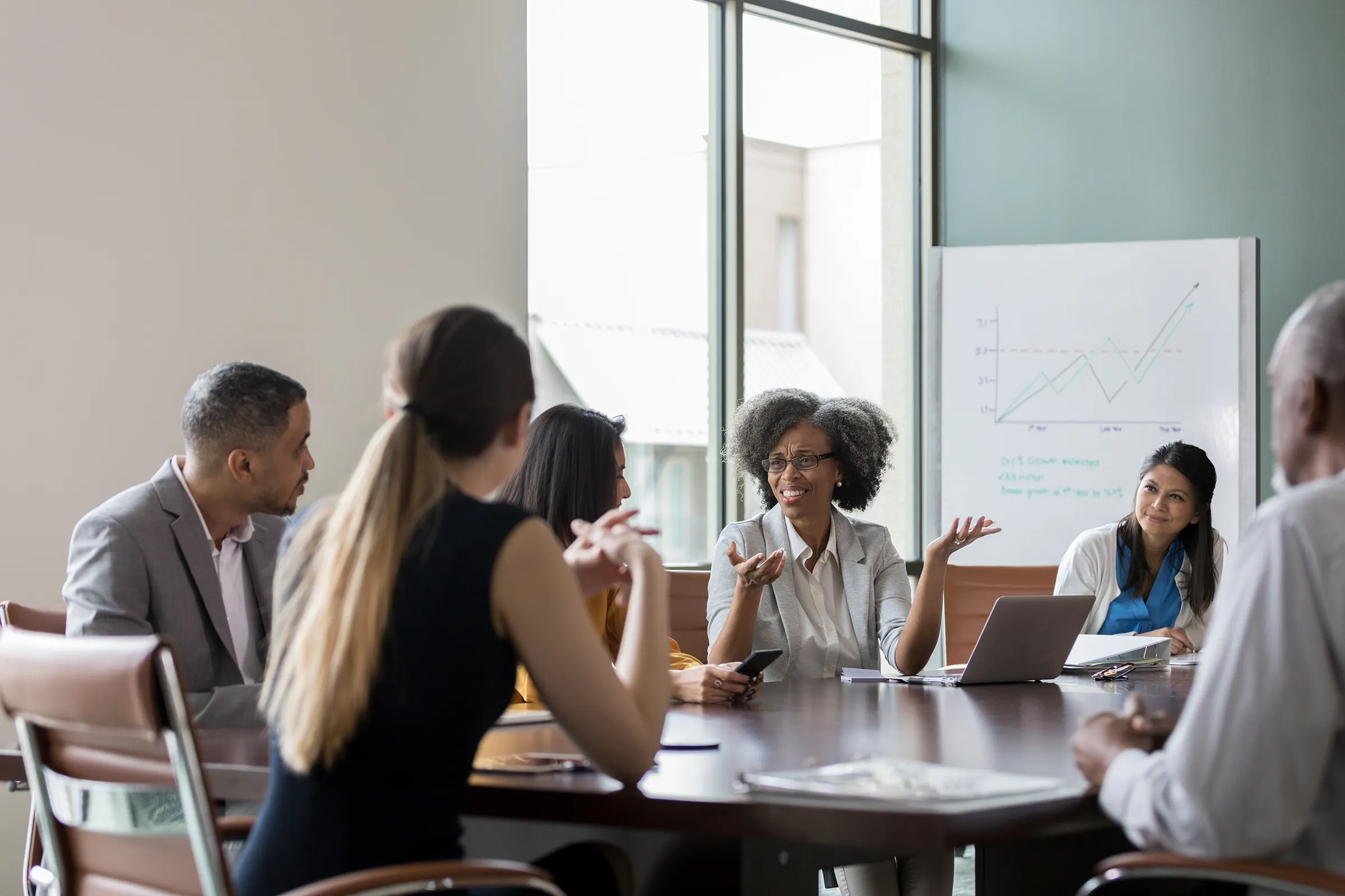 A woman with glasses gestures as she speaks during a meeting around a conference table, with colleagues listening and a line chart on a whiteboard behind her