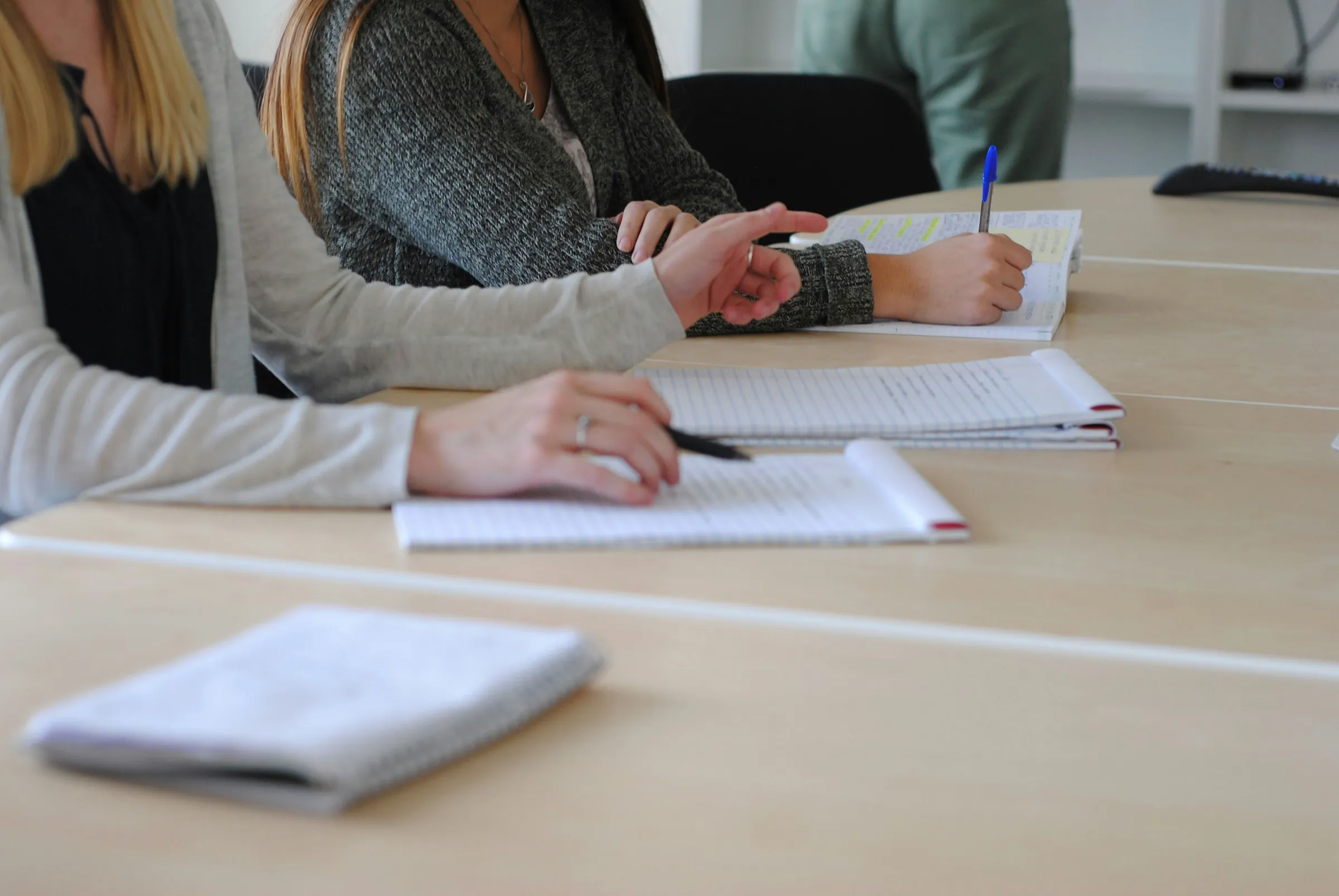 Close-up of two people seated at a conference table taking handwritten notes on notepads