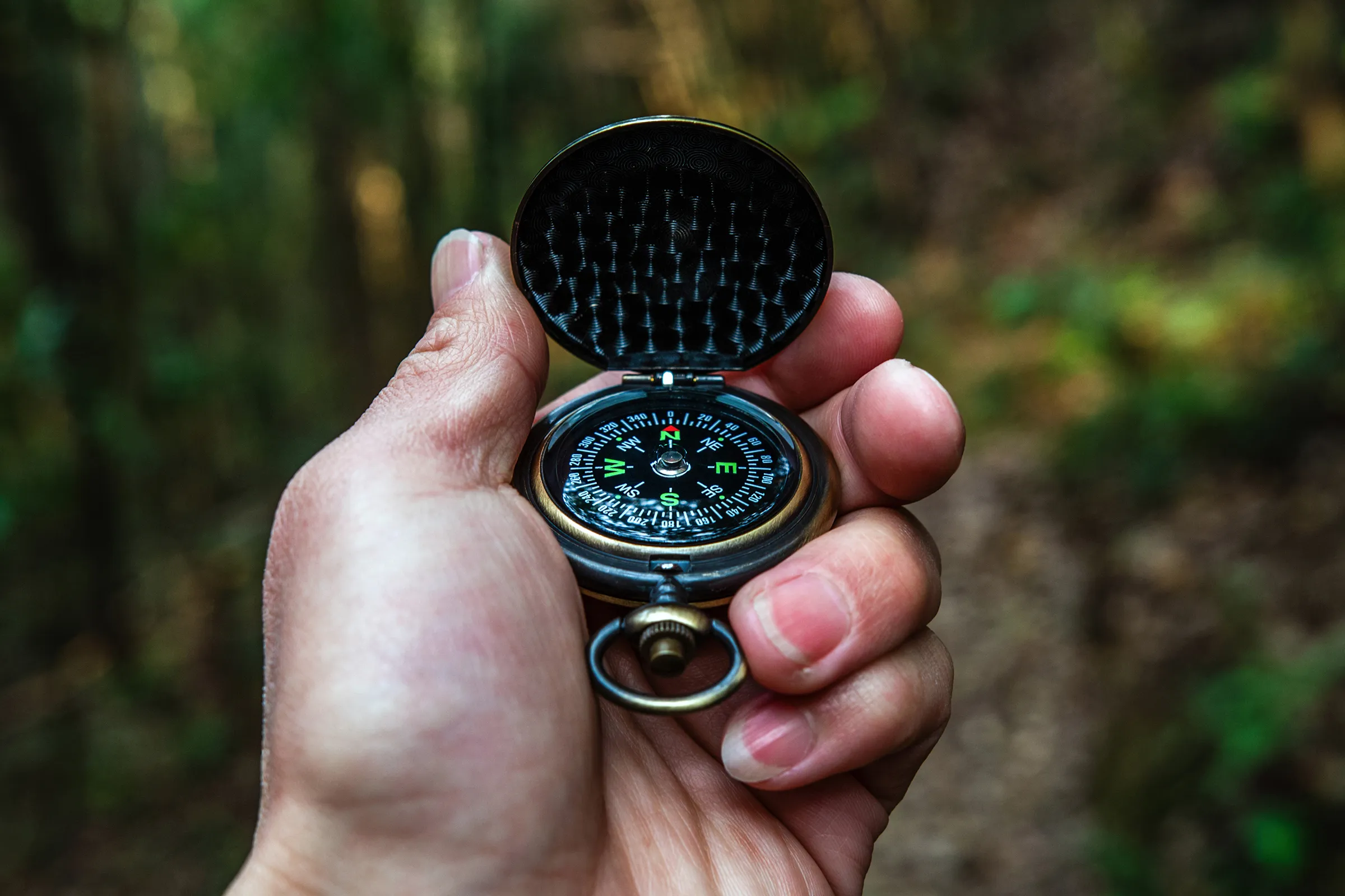 A hand holding an open brass pocket compass on a wooded trail
